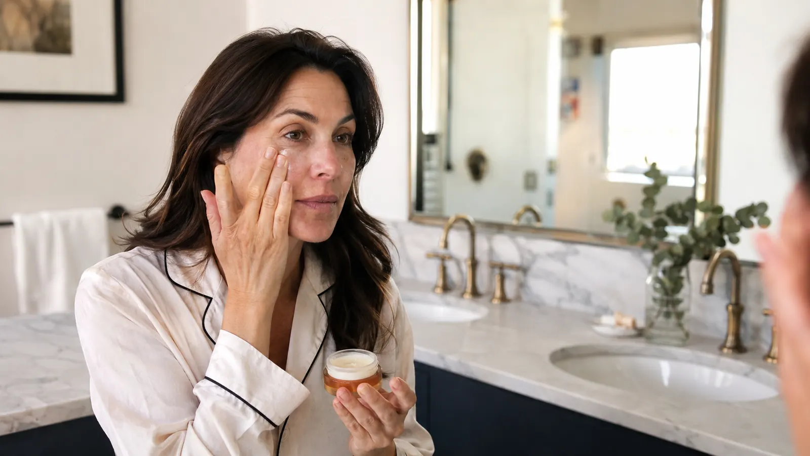 Woman gently applying eye cream under her eyes at a bathroom vanity with warm morning light